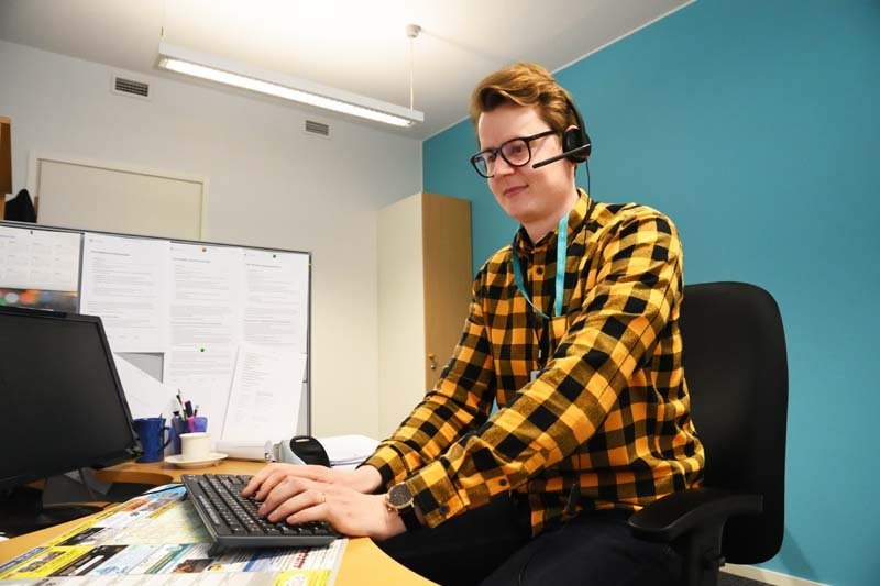 Asiakasneuvoja Jaakko Puputti istuu työpisteellään. Hänellä on luurit päässä ja hän näppäilee tietokoneen näppäimistöä. Huoneessa on turkoosinsininen seinä.