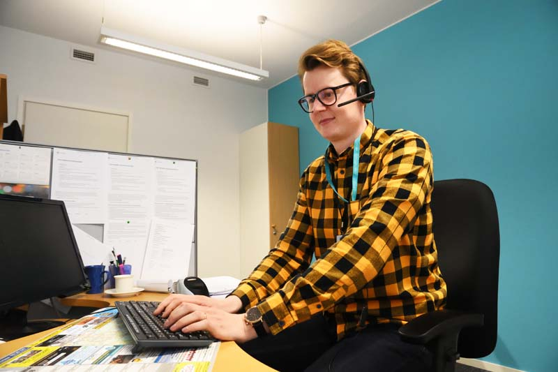 Asiakasneuvoja Jaakko Puputti istuu työpisteellään. Hänellä on luurit päässä ja hän näppäilee tietokoneen näppäimistöä. Huoneessa on turkoosinsininen seinä.