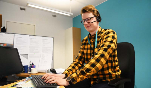 Asiakasneuvoja Jaakko Puputti istuu työpisteellään. Hänellä on luurit päässä ja hän näppäilee tietokoneen näppäimistöä. Huoneessa on turkoosinsininen seinä.