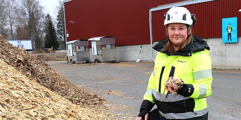 Operativa chefen Karoliina Kärkäs står bredvid en flishög i arbetskläder och med hjälm på huvudet. I bakgrunden syns en stor byggnad.