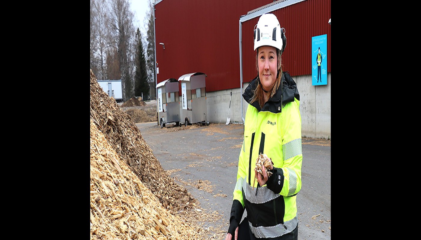 Operativa chefen Karoliina Kärkäs står bredvid en flishög i arbetskläder och med hjälm på huvudet. I bakgrunden syns en stor byggnad.