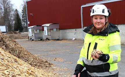 Operativa chefen Karoliina Kärkäs står bredvid en flishög i arbetskläder och med hjälm på huvudet. I bakgrunden syns en stor byggnad.