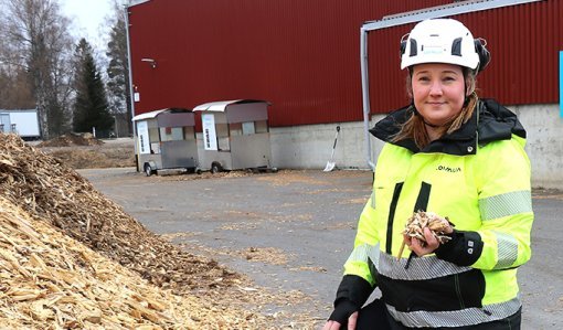 Operativa chefen Karoliina Kärkäs står bredvid en flishög i arbetskläder och med hjälm på huvudet. I bakgrunden syns en stor byggnad.