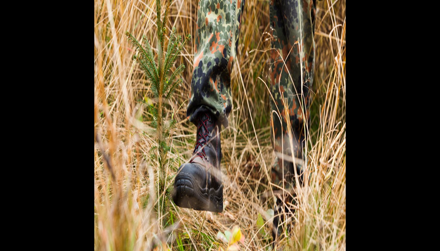 Lähikuva maastokengistä, jotka polkevat ruskeaa heinää matalaksi taimen ympäriltä. 