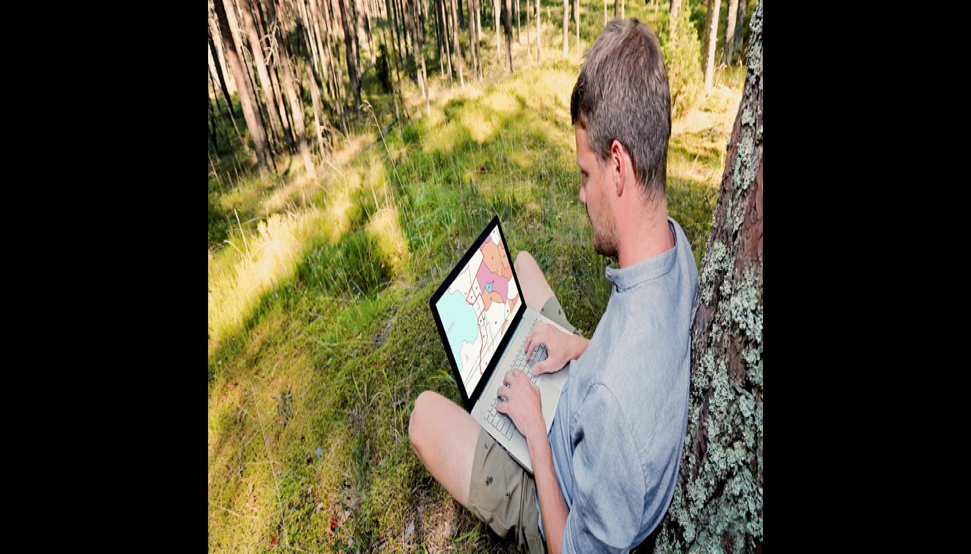 Mies istuu nurmikolla kannettava tietokone sylissään ja katsoo oman metsänsä tietoja Metsään.fi-palvelusta. Hän nojaa puuhun. Miehen ympärillä on nurmikkoa ja taustalla on metsää.