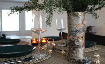 Jouluinen kattaus. Pöydässä lautaset, aterimet, viinilasit, kaksi tuikkukynttilää ja koristepalloja ja -käpyjä. Edessä koivumaljakko, jossa havunoksia.