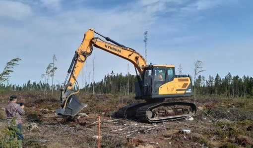 Metsäkylvökauha on kiinnitetty kaivinkoneeseen ja tekee kylvöä hakkuuaukealla. Vasemmalla henkilö ottaa kuvaa koneesta.