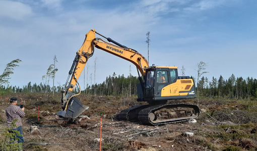 Metsäkylvökauha on kiinnitetty kaivinkoneeseen ja tekee kylvöä hakkuuaukealla. Vasemmalla henkilö ottaa kuvaa koneesta.