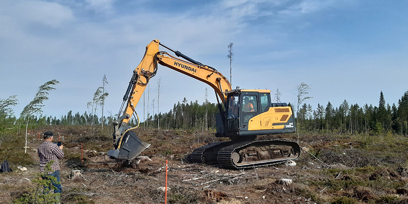 Metsäkylvökauha on kiinnitetty kaivinkoneeseen ja tekee kylvöä hakkuuaukealla. Vasemmalla henkilö ottaa kuvaa koneesta.