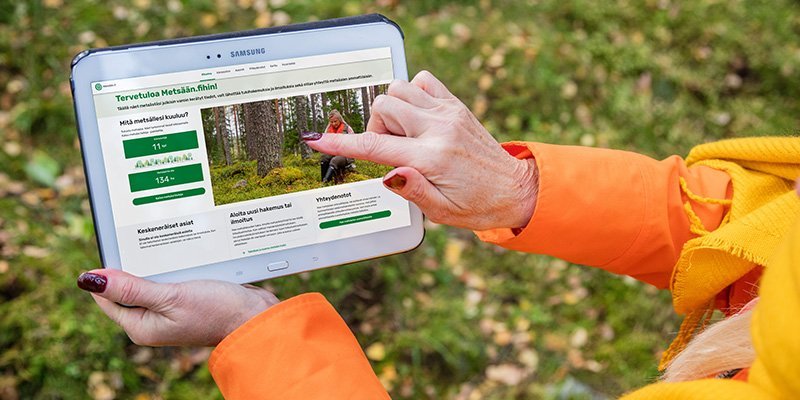 Henkilö pitää kädessään tablettia, jossa on avoinna Metsään.fi-palvelun etusivu. Hän osoittaa toisella sormellaan etusivua. 