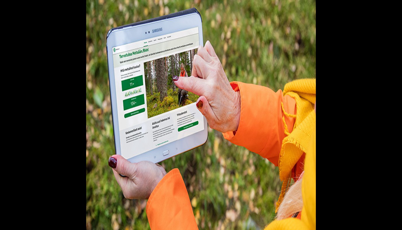 Henkilö pitää kädessään tablettia, jossa on avoinna Metsään.fi-palvelun etusivu. Hän osoittaa toisella sormellaan etusivua. 