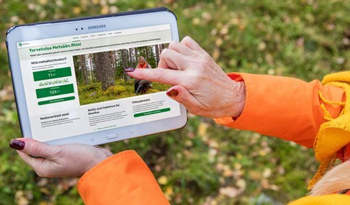 Henkilö pitää kädessään tablettia, jossa on avoinna Metsään.fi-palvelun etusivu. Hän osoittaa toisella sormellaan etusivua. 