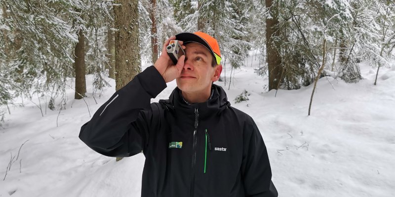 Metsäneuvoja Ari Kasanen katsoo ja mittailee korkeusmittarilla puustoa talvisessa metsässä. 