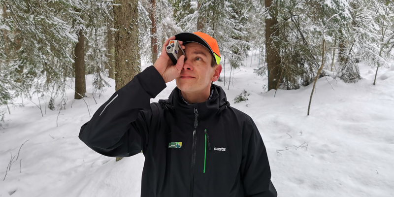Metsäneuvoja Ari Kasanen katsoo ja mittailee korkeusmittarilla puustoa talvisessa metsässä.