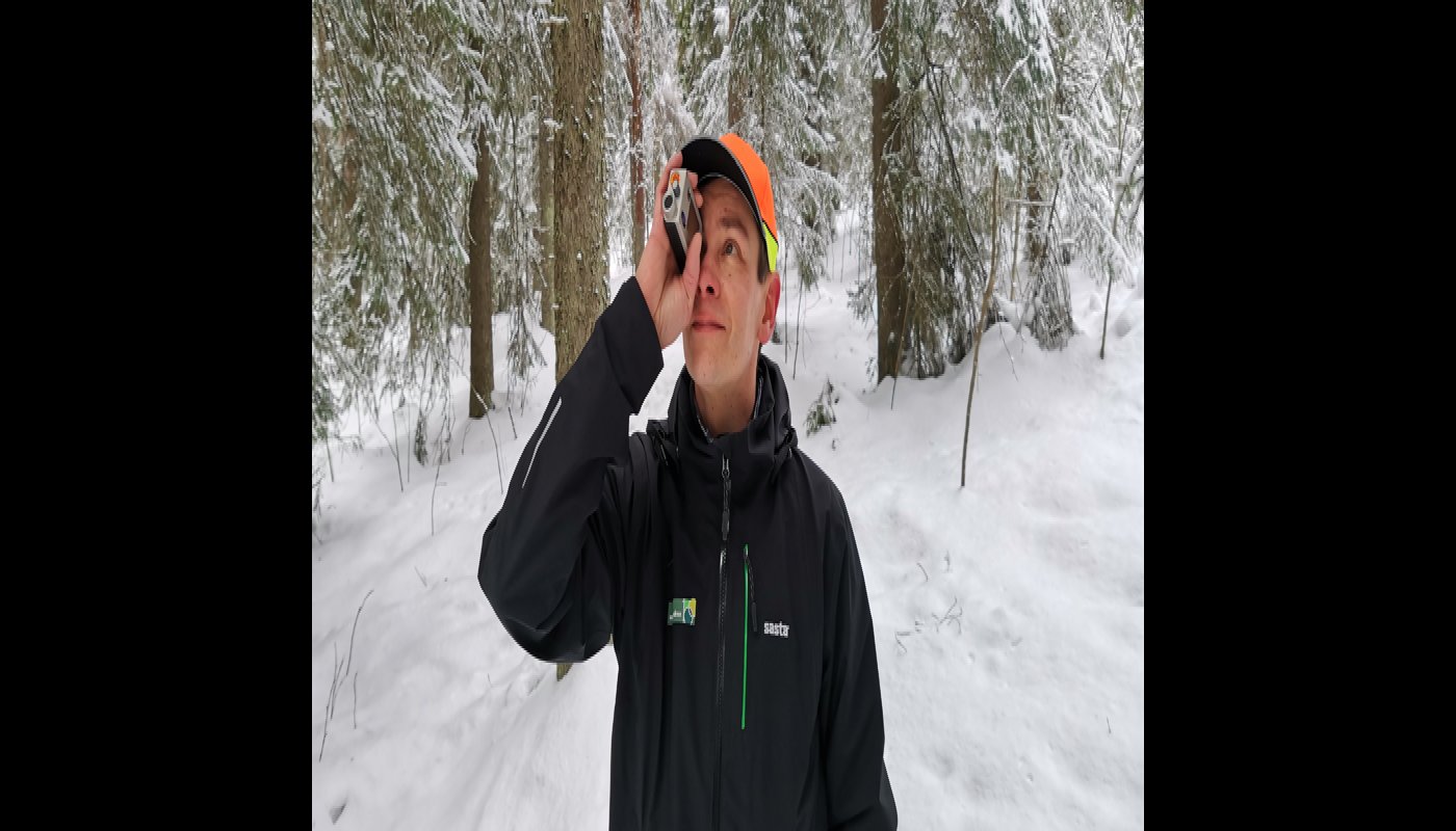 Metsäneuvoja Ari Kasanen katsoo ja mittailee korkeusmittarilla puustoa talvisessa metsässä. 