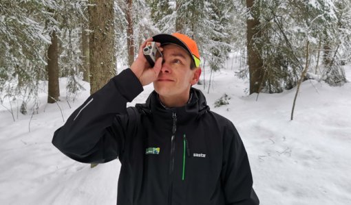 Metsäneuvoja Ari Kasanen katsoo ja mittailee korkeusmittarilla puustoa talvisessa metsässä. 