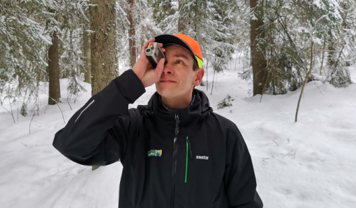 Metsäneuvoja Ari Kasanen katsoo ja mittailee korkeusmittarilla puustoa talvisessa metsässä.