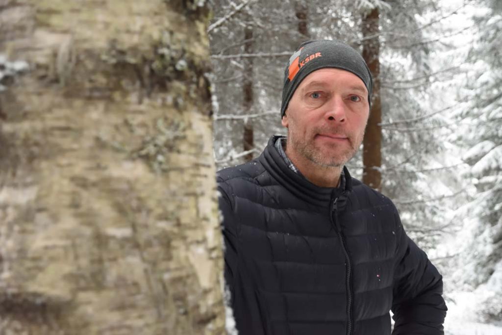 Mustaan toppatakkiin pukeutunut Arto Minkkinen kurkistaa paksun koivunrungon takaa. Taustalla on lumista metsää.