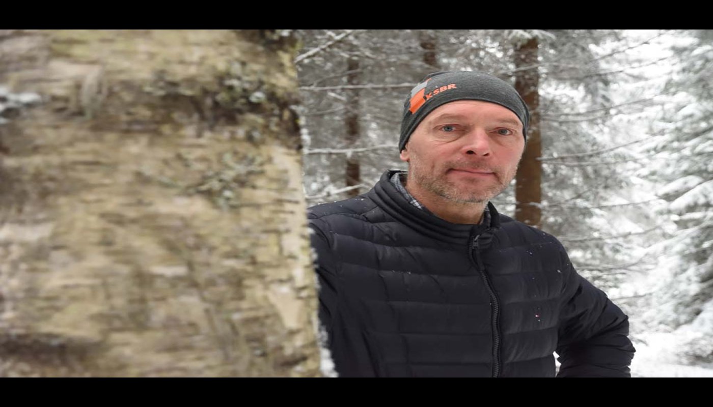 Mustaan toppatakkiin pukeutunut Arto Minkkinen kurkistaa paksun koivunrungon takaa. Taustalla on lumista metsää.