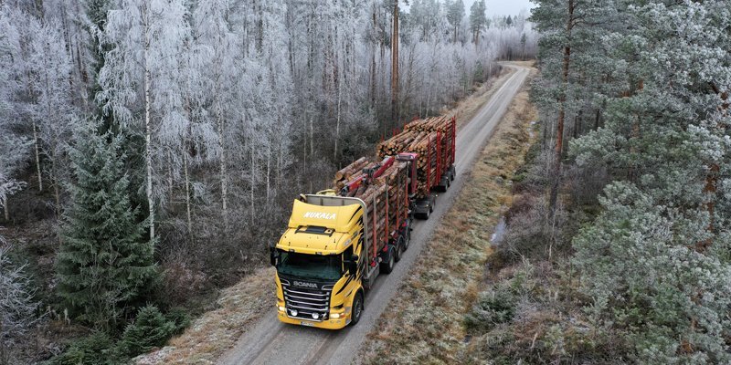 Tukkirekka kuvattuna ylhäältäpäin. Rekassa on keltainen ohjaamo ja se on täydessä kuormassa. Metsätien ympärillä on kuuraisia puita.