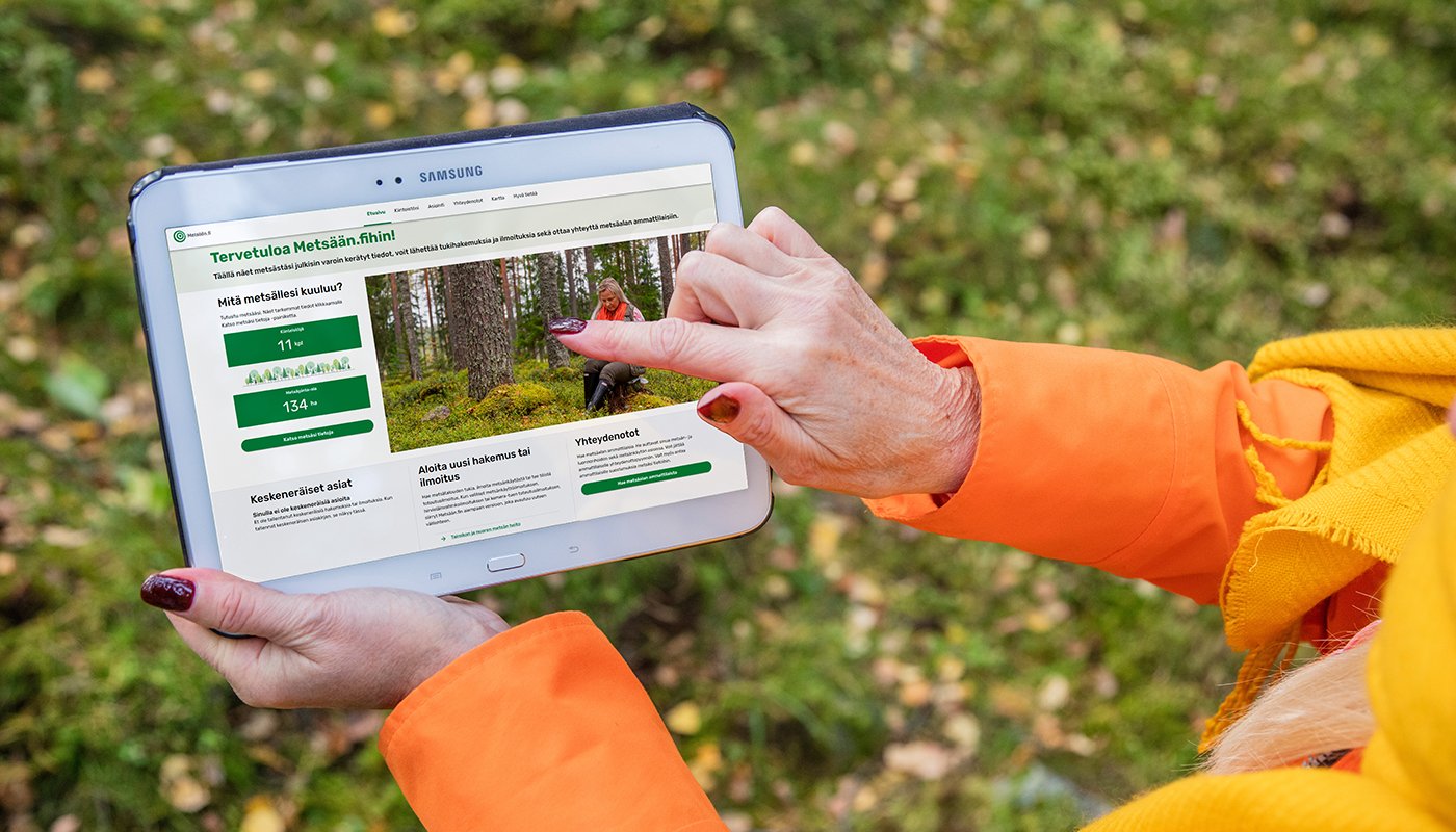 Henkilö pitää kädessään tablettia, jossa on avoinna Metsään.fi-palvelun etusivu. 
