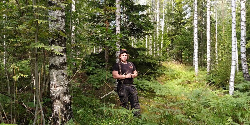 Tuomas Korpijaakko seisoo keskellä kuvaa omassa metsässään, jossa kasvaa sekapuustoa. Kuvassa hänen ympärillään on koivuja, pihlajia, kuusia ja saniaisia. 