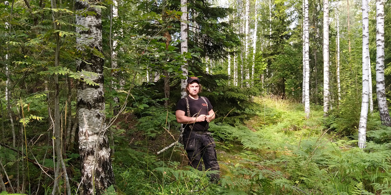Tuomas Korpijaakko seisoo keskellä kuvaa omassa metsässään, jossa kasvaa sekapuustoa. Kuvassa hänen ympärillään on koivuja, pihlajia, kuusia ja saniaisia.