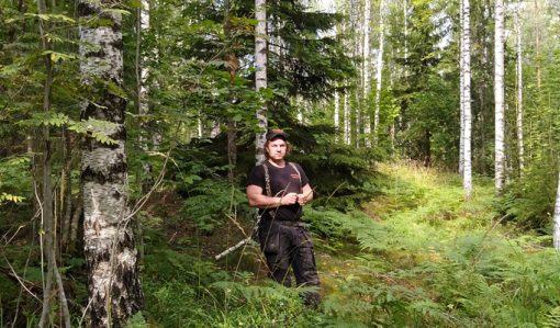 Tuomas Korpijaakko seisoo keskellä kuvaa omassa metsässään, jossa kasvaa sekapuustoa. Kuvassa hänen ympärillään on koivuja, pihlajia, kuusia ja saniaisia.
