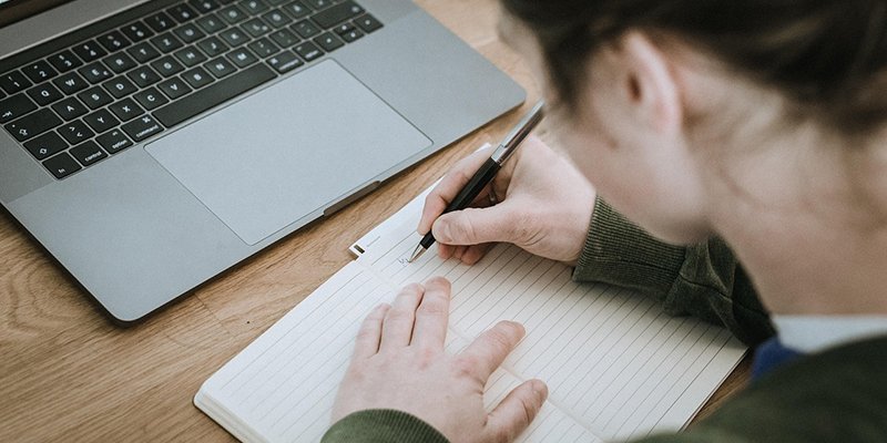 Henkilö kirjoittaa muistiinpanoja vihkoon. Vieressä pöydällä on kannettava tietokone.
