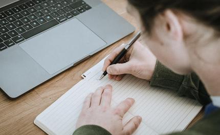 Henkilö kirjoittaa muistiinpanoja vihkoon. Vieressä pöydällä on kannettava tietokone.
