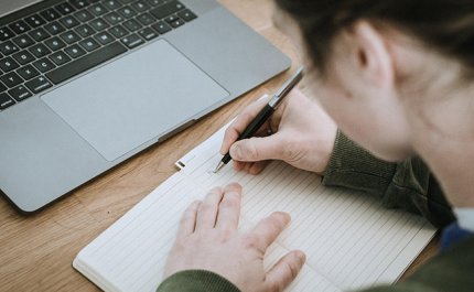 Henkilö kirjoittaa muistiinpanoja vihkoon. Vieressä pöydällä on kannettava tietokone.
