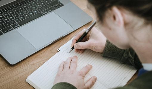 Henkilö kirjoittaa muistiinpanoja vihkoon. Vieressä pöydällä on kannettava tietokone.

