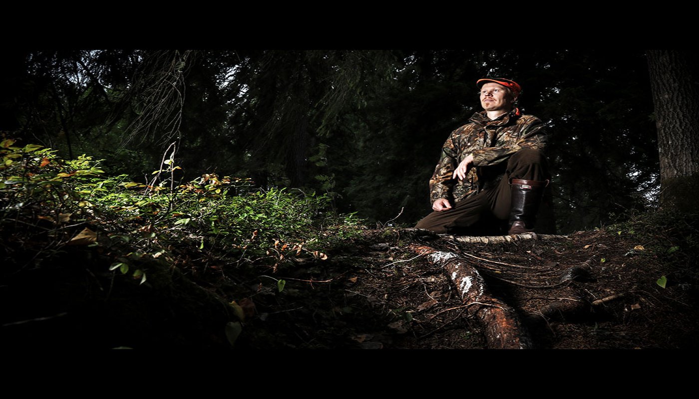 Riistasuunnittelija Teemu Lamberg on nostanut jalkansa ison puun juurakon päälle. Ympärillä on tummasävyistä metsää ja maassa varvikkoa. 