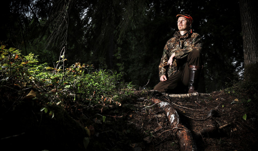 Riistasuunnittelija Teemu Lamberg on nostanut jalkansa ison puun juurakon päälle. Ympärillä on tummasävyistä metsää ja maassa varvikkoa.