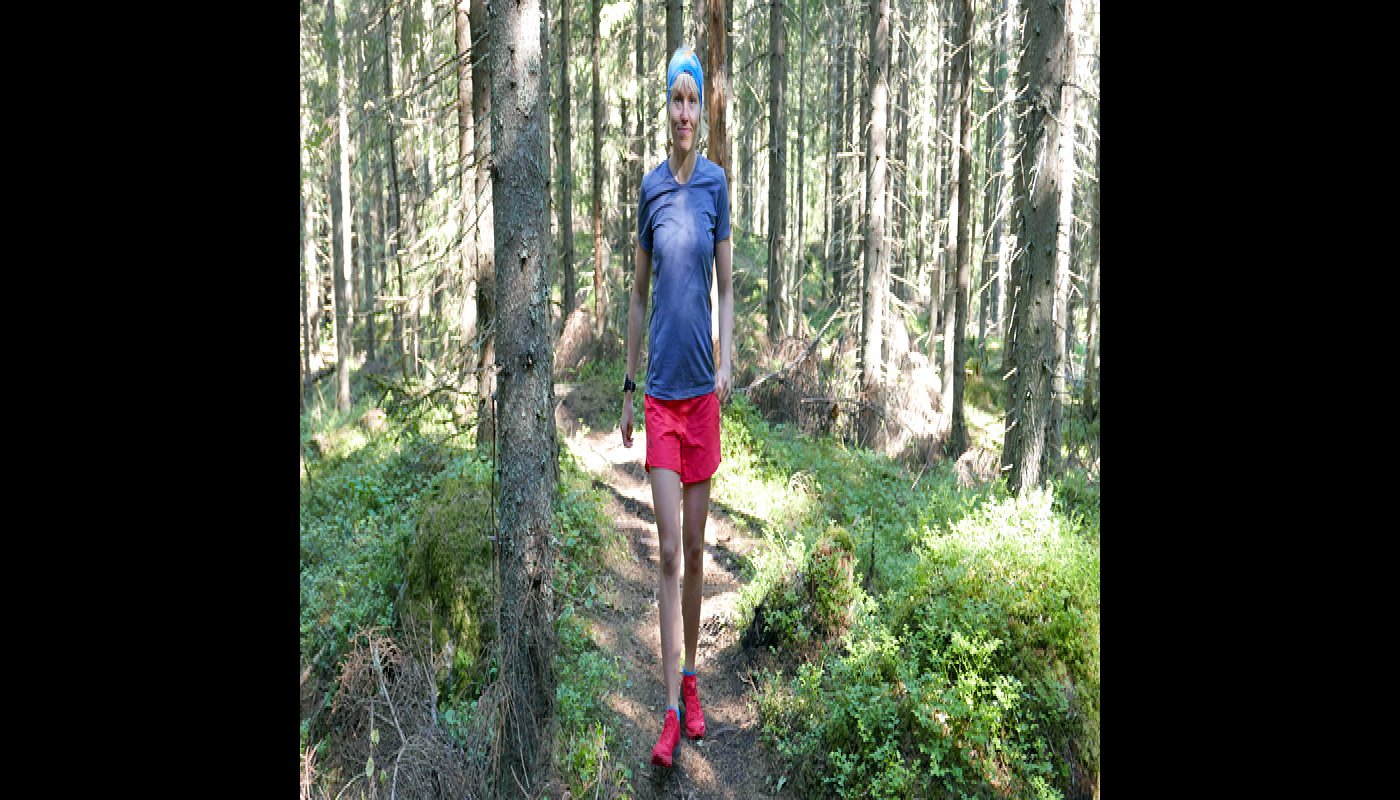 Saara Grönholm kävelee eteenpäin polulla ja hymyilee. Ympärillä on kuusimetsää ja varvikkoa. 