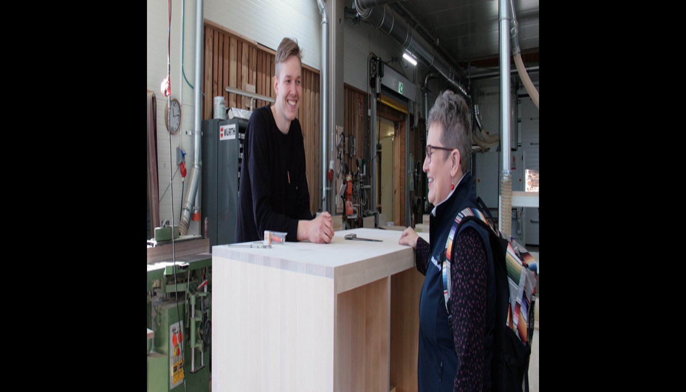 Miikka Kotilainen juttelee puisen pöytätason takaa yritysneuvoja Leila Laukkaselle Puuartistin tuotantotiloissa. 