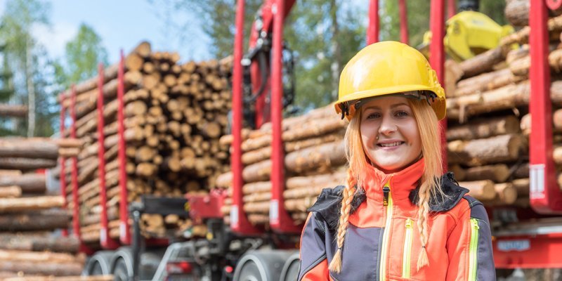 Sarah Lindahl keltainen turvakypärä päässään on kuvan etualalla, taustalla erottuu lähes täydessä lastissa oleva puunkuljetusauto. 