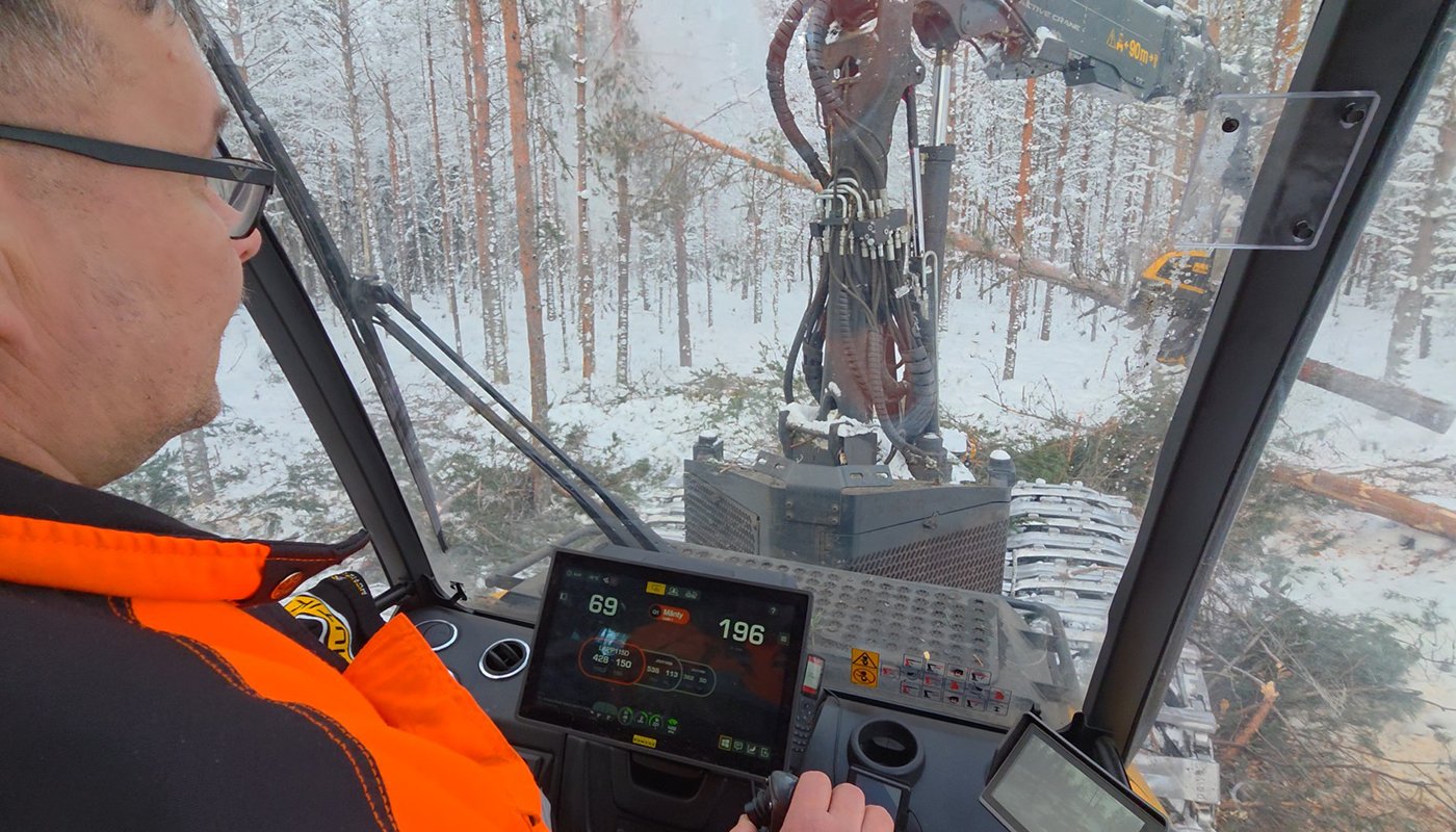 Hakkuukoneen ohjaamosta kuvaa. Kone kaataa puuta talvisessa metsässä.  
