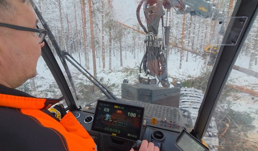 Hakkuukoneen ohjaamosta kuvaa. Kone kaataa puuta talvisessa metsässä.  
