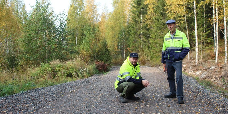 Tieisännöitsijä Tomi Vähä-Kouvola on kyykyssä metsätiellä ja hänen vieressään seisoo Tapio Kahilaniemi. Tielle on pudonnut keltaisia lehtiä ja tien ympärillä on metsää.