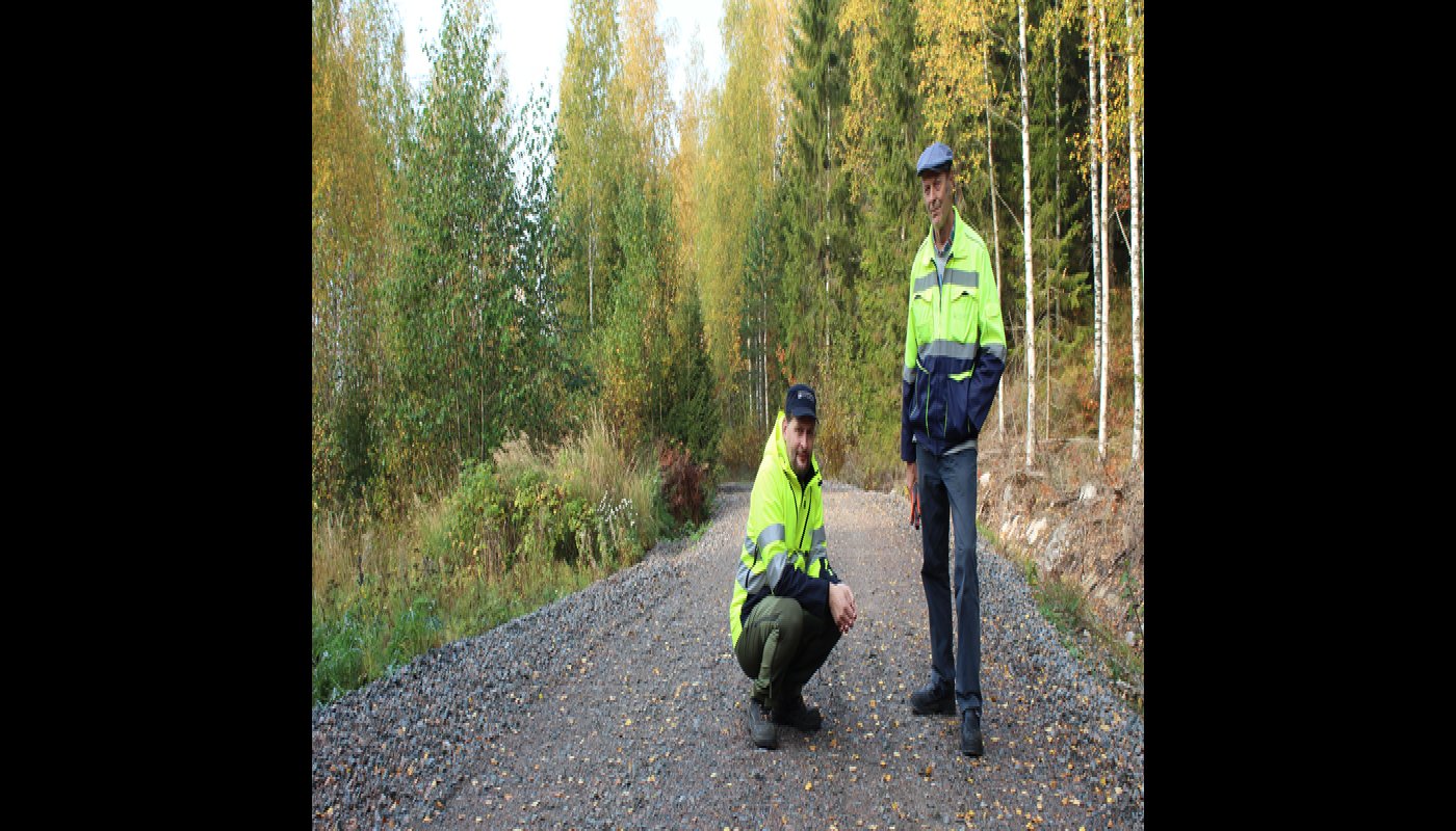 Tieisännöitsijä Tomi Vähä-Kouvola on kyykyssä metsätiellä ja hänen vieressään seisoo Tapio Kahilaniemi. Tielle on pudonnut keltaisia lehtiä ja tien ympärillä on metsää.