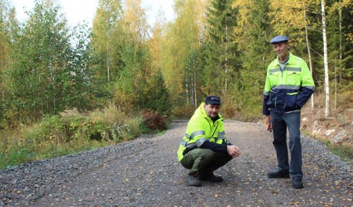 Tieisännöitsijä Tomi Vähä-Kouvola on kyykyssä metsätiellä ja hänen vieressään seisoo Tapio Kahilaniemi. Tielle on pudonnut keltaisia lehtiä ja tien ympärillä on metsää.