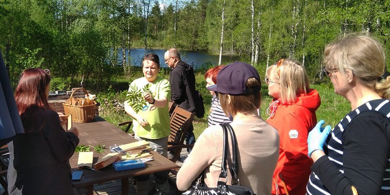 Luonnonvaratuottaja Leena Löf esittelee villiyrttejä ympärillään oleville kuulijoille. Taustalla on koivuja ja järvimaisemaa.