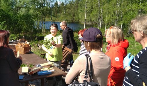 Luonnonvaratuottaja Leena Löf esittelee villiyrttejä ympärillään oleville kuulijoille. Taustalla on koivuja ja järvimaisemaa.