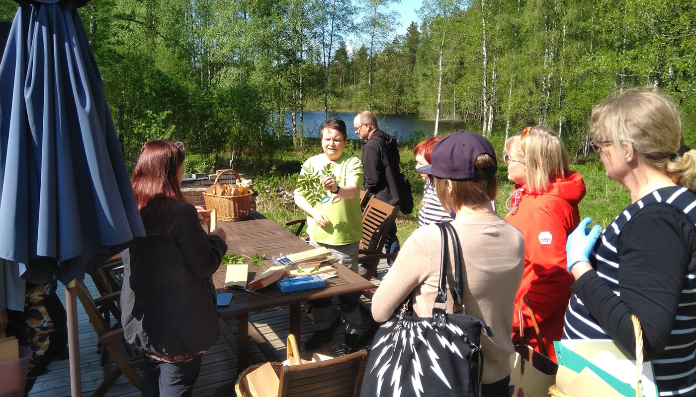Luonnonvaratuottaja Leena Löf esittelee villiyrttejä ympärillään oleville kuulijoille. Taustalla on koivuja ja järvimaisemaa.