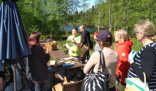 Luonnonvaratuottaja Leena Löf esittelee villiyrttejä ympärillään oleville kuulijoille. Taustalla on koivuja ja järvimaisemaa.