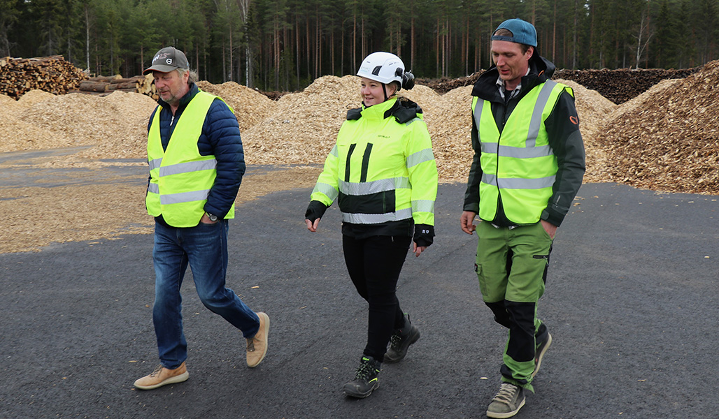 Kuljetusliikkeen yrittäjä Jarmo Hieto (vas.), Loimua Oy:n operointipäällikkö Karoliina Kärkäs sekä maa- ja metsätalousyrittäjä Lauri Tapola Hiedon puuterminaalilla. Hieto ja Tapola ovat pukeutuneet keltaisiin huomioliiveihin, Kärkkäällä on keltainen työmaatakki. Taustalla on hake- ja puukasoja.