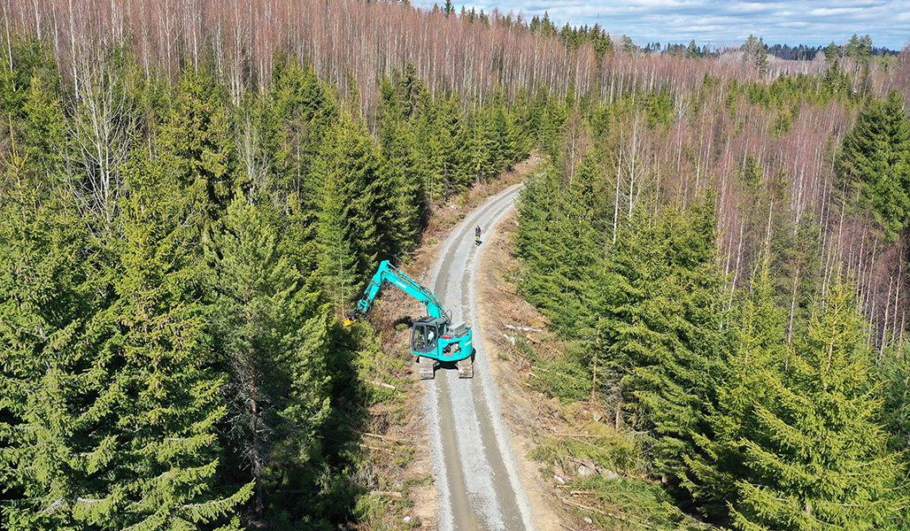 Ylhäältä päin otetussa kuvassa metsätietä pitkin kulkee kaivinkone, jonka giljotiinipää leikkaa puustoa tien reunasta. Tiellä kauempana työtä seuraa ihminen. Tien ympärillä on kuusikkoa ja koivikkoa.