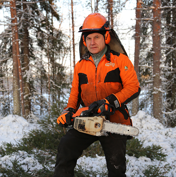 Alpo Särkelä oranssimustissa turvavarusteissa moottorisahan kanssa talvisessa metsässä. 