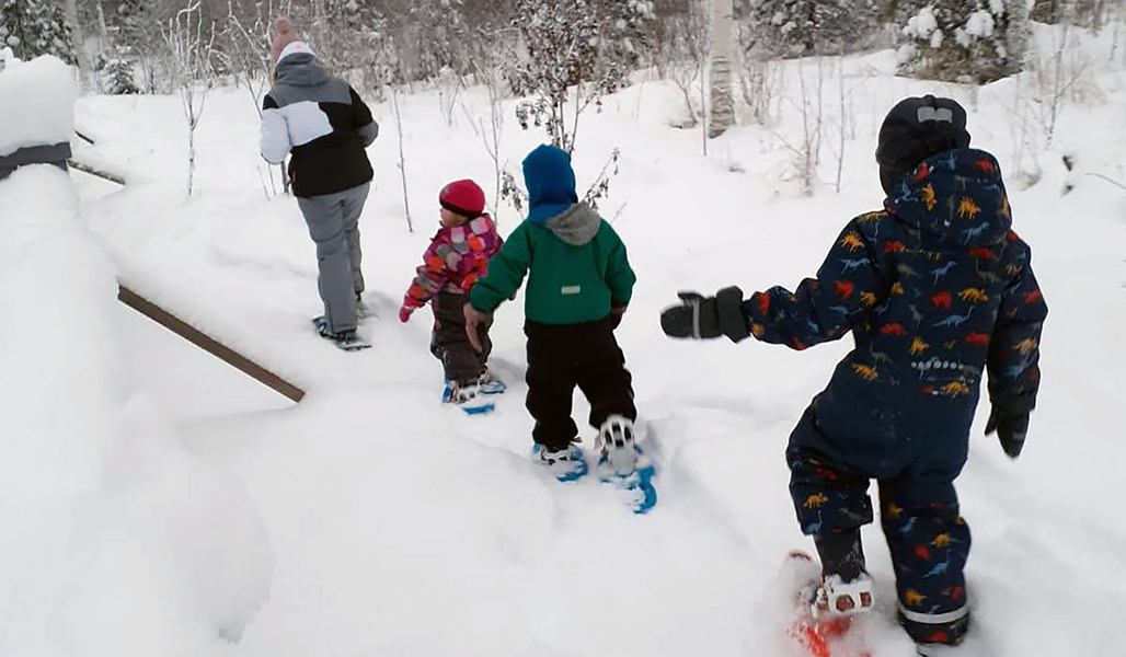 Aikuinen ja kolme lasta kulkevat hangessa lumikengillä talvella. 